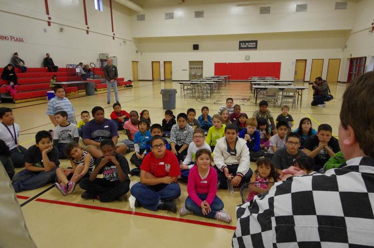 Tournament participants receive a chess lesson from Professional Chess Teacher Ms. Daa (right, in chess-squares shirt). In the background, the competition tables await.